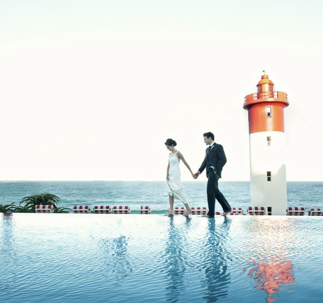 Brautpaar spaziert am Infinity-Pool mit rotem Leuchtturm im Hintergrund – Destination Wedding in Südafrika – Hochzeitsplanerin Essen
